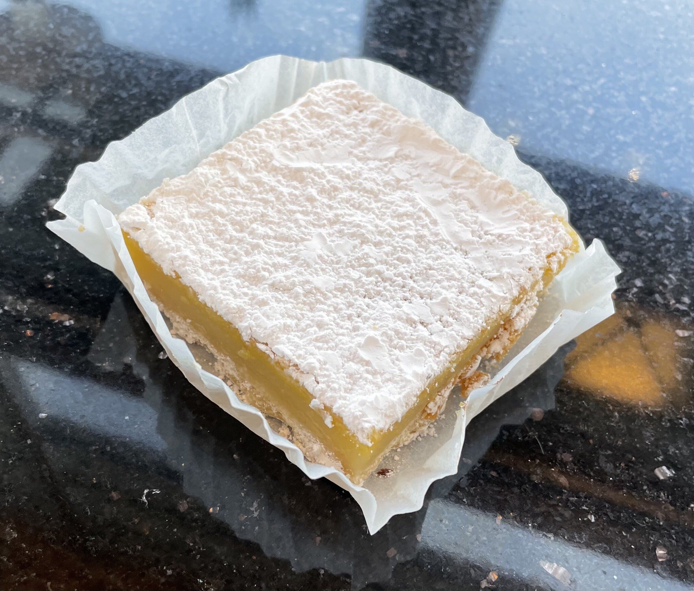 A lemon bar dusted with powdered sugar in a paper liner